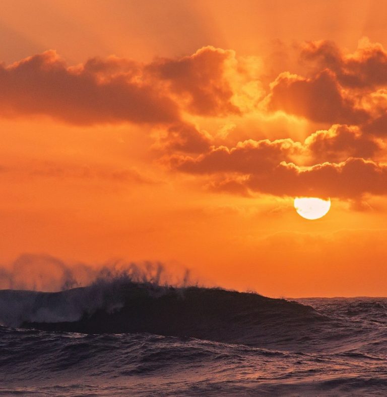 Sunset Waves Dramatic orange sunset sky with sun visible through clouds over silhouetted breaking waves.